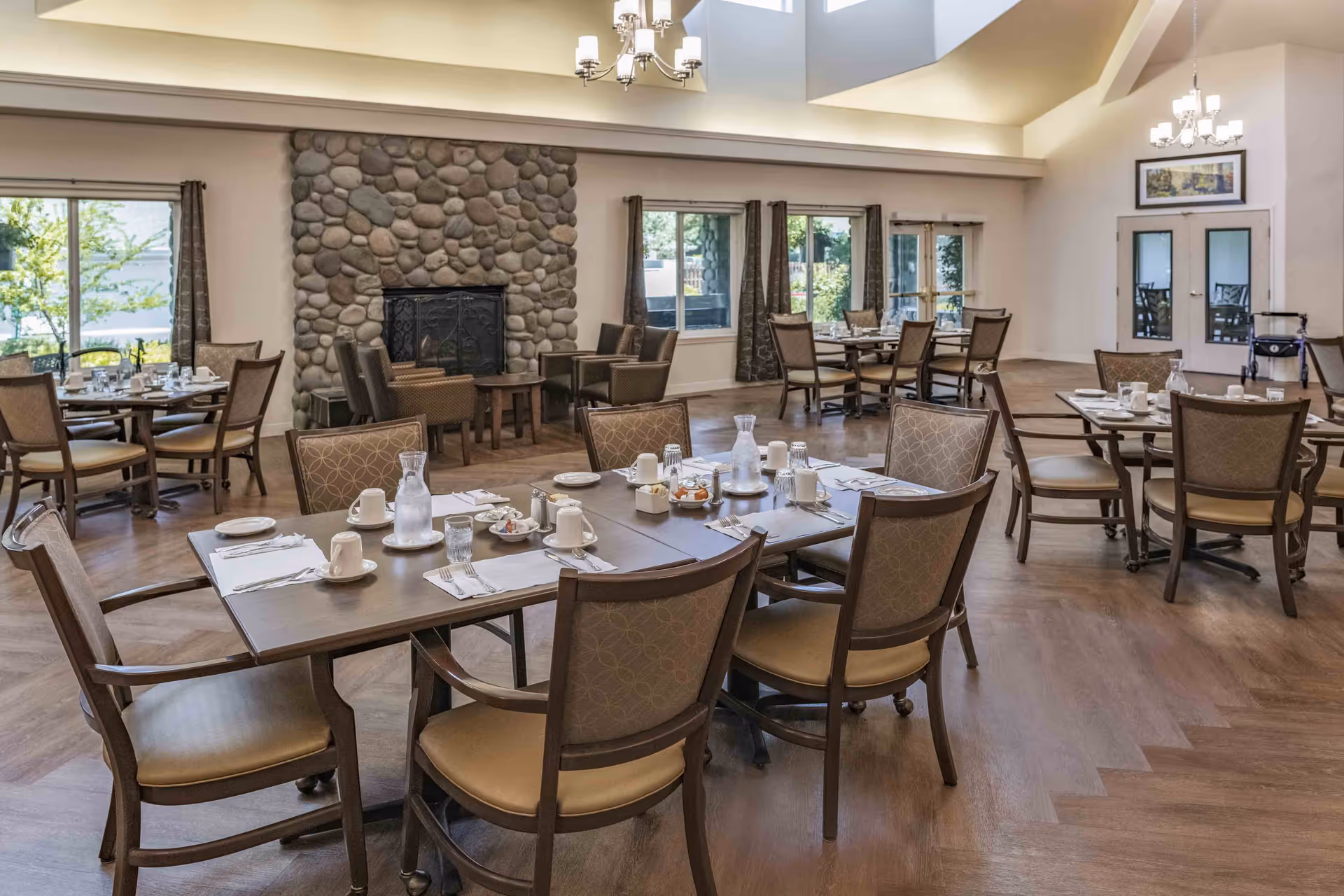 A spacious dining room with multiple tables and chairs arranged neatly. Each table is set with white cups, plates, napkins, and glass water pitchers. The room features large windows with curtains, a stone fireplace, wooden flooring, and chandeliers hanging from the ceiling. There is a walker near the double doors at the back.