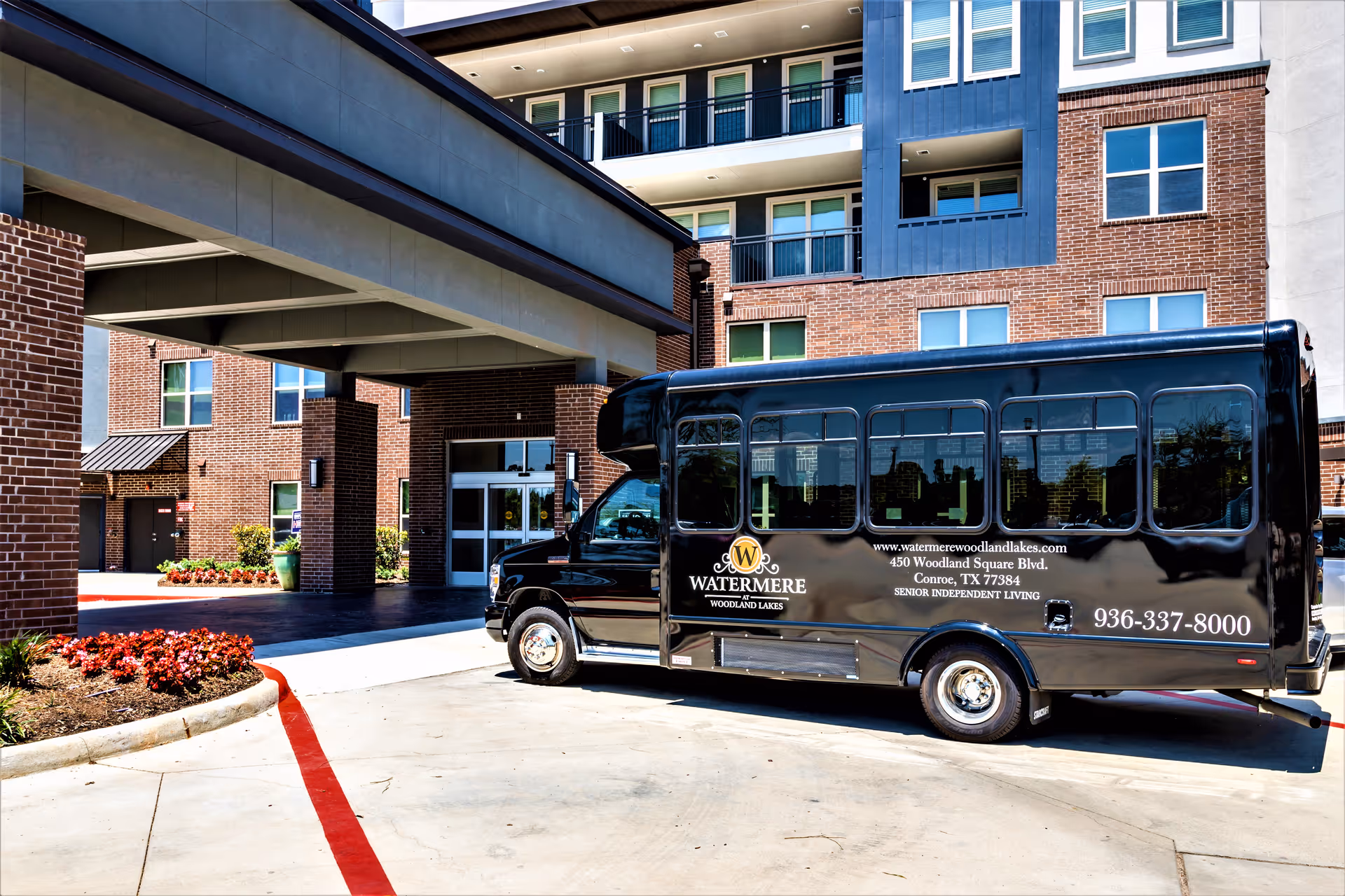 A black shuttle bus parked in front of the entrance to a multi-story brick building with balconies. The bus has the Watermere at Woodland Lakes logo and contact information on its side. The building entrance is covered by a large overhang supported by brick pillars, and there are landscaped flower beds nearby.