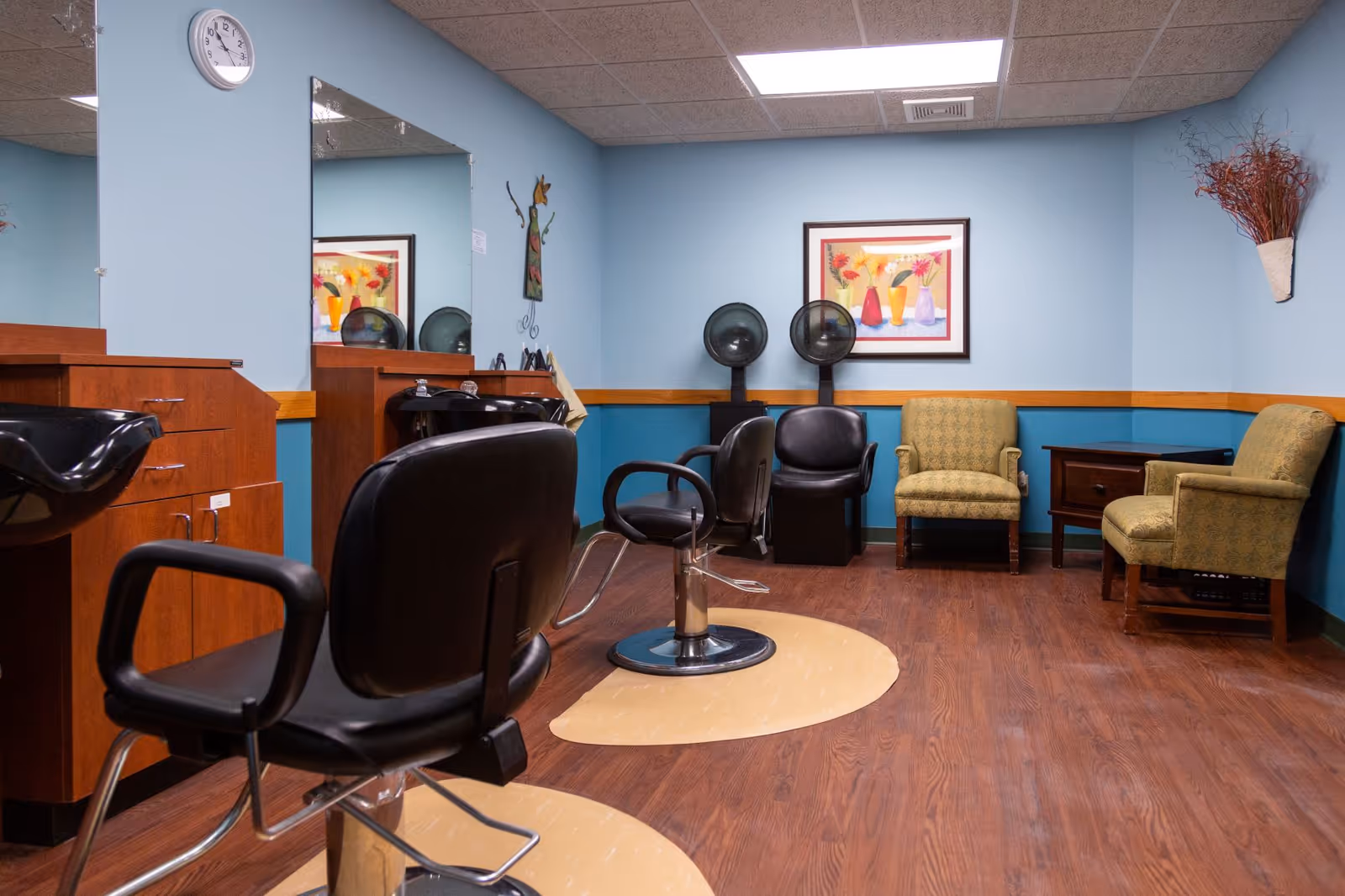 Interior of a salon area in a senior living facility with two black salon chairs and hair washing stations on the left, two hair dryers against the back wall, two upholstered armchairs, a small wooden table, and colorful artwork on the blue walls.