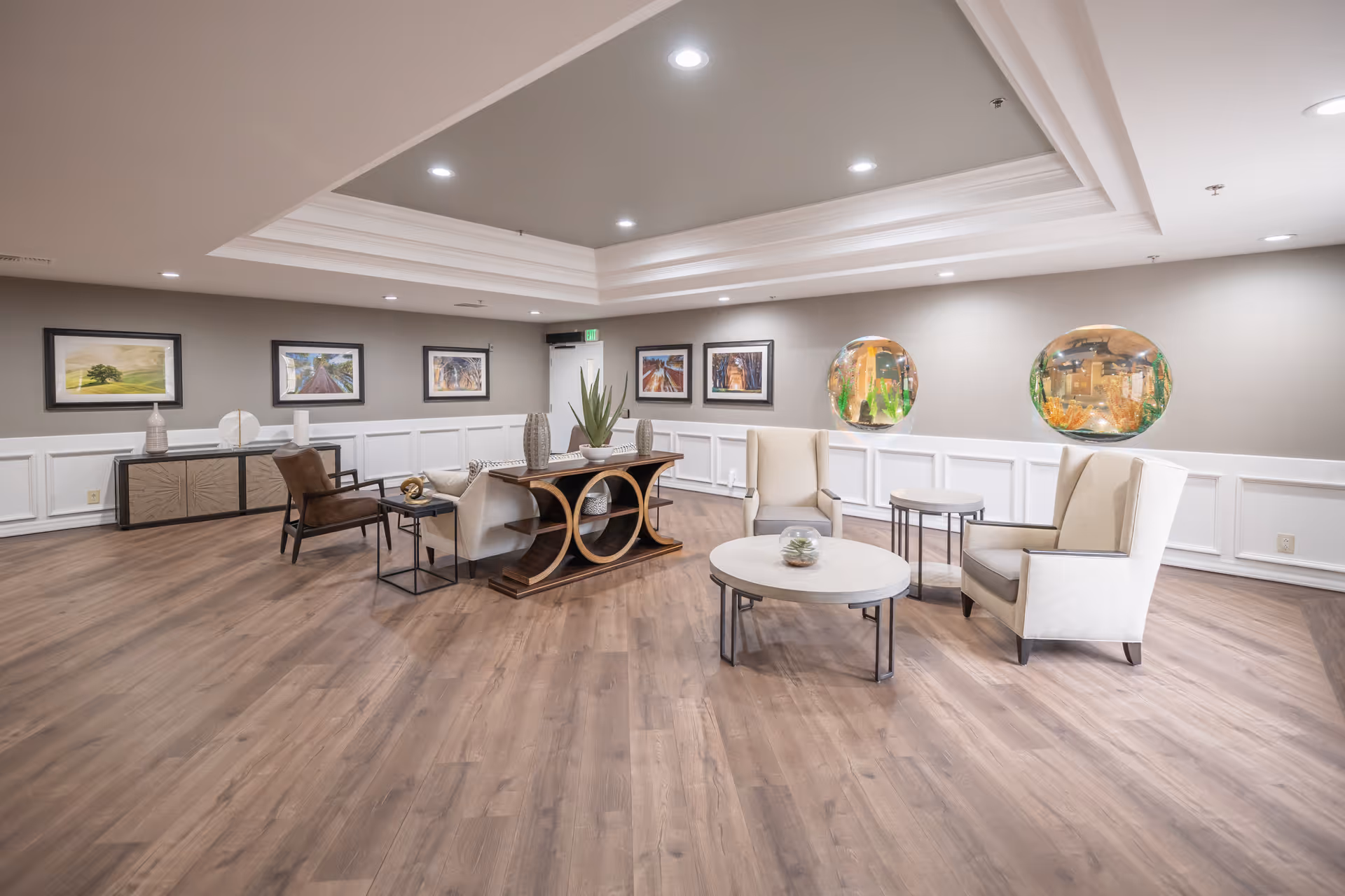 A spacious, well-lit lounge area with modern furniture including beige armchairs, a sofa, and round coffee tables. The room features wood flooring, gray walls with white wainscoting, framed artwork, and two large round wall-mounted aquariums. The ceiling has recessed lighting and a tray design.