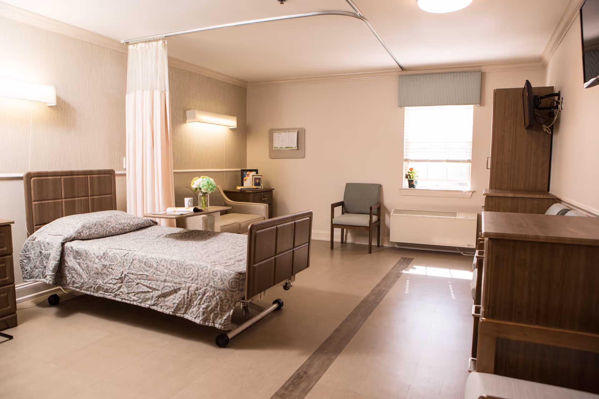 A well-lit senior living bedroom featuring a single hospital-style bed with patterned bedding, a privacy curtain, a small table with a vase of flowers, an armchair, a window with blinds, a wall-mounted TV, and wooden furniture including a dresser and a cabinet.
