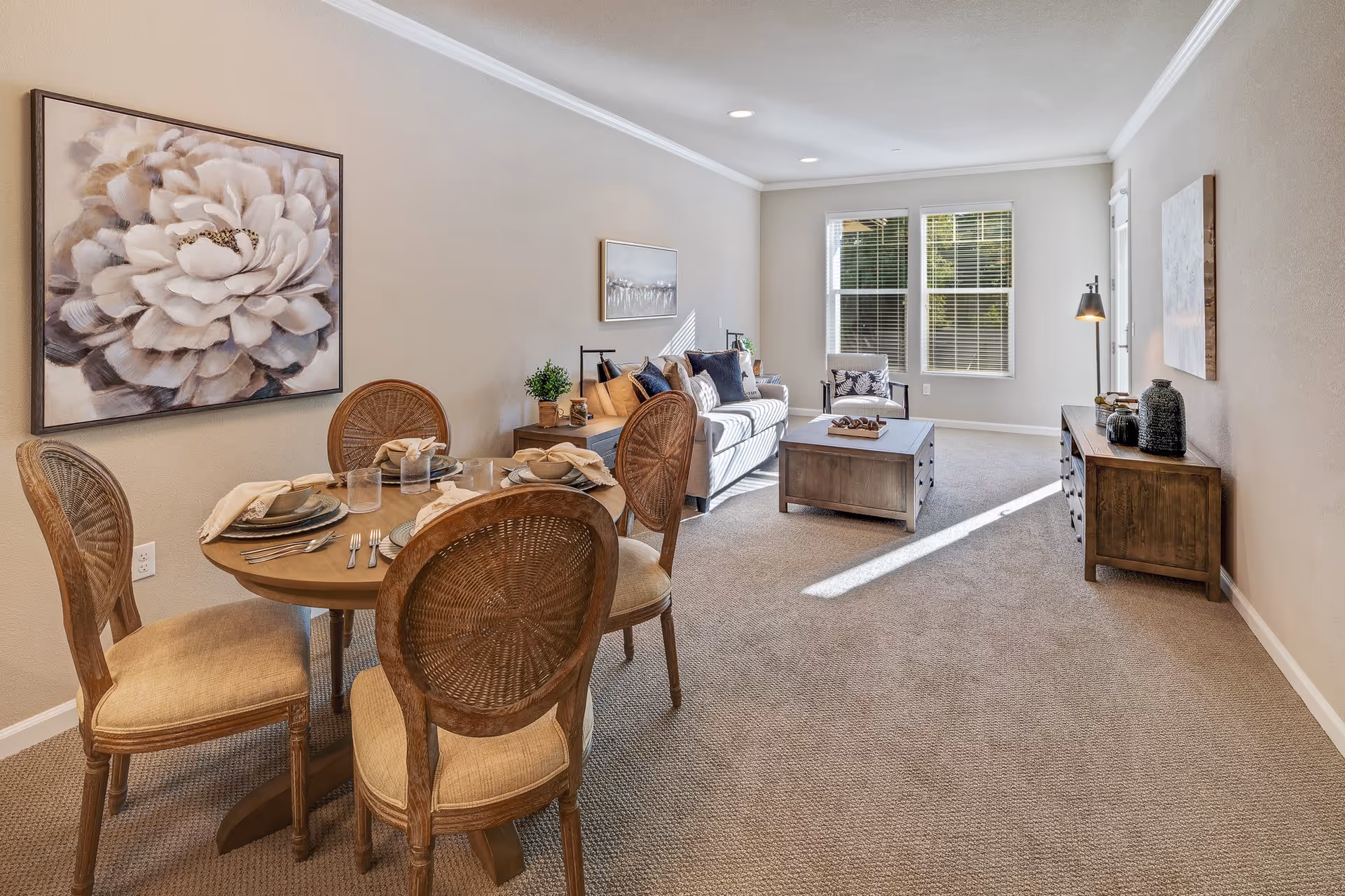 A well-lit living and dining area featuring a round wooden dining table set with four chairs, each with woven backs and cushioned seats. The table is set with plates, napkins, and glasses. In the background, there is a cozy living room with a sofa, armchair, coffee table, and a wooden sideboard. The walls are painted beige, adorned with large floral and landscape artwork. Sunlight streams in through two large windows, illuminating the carpeted floor.