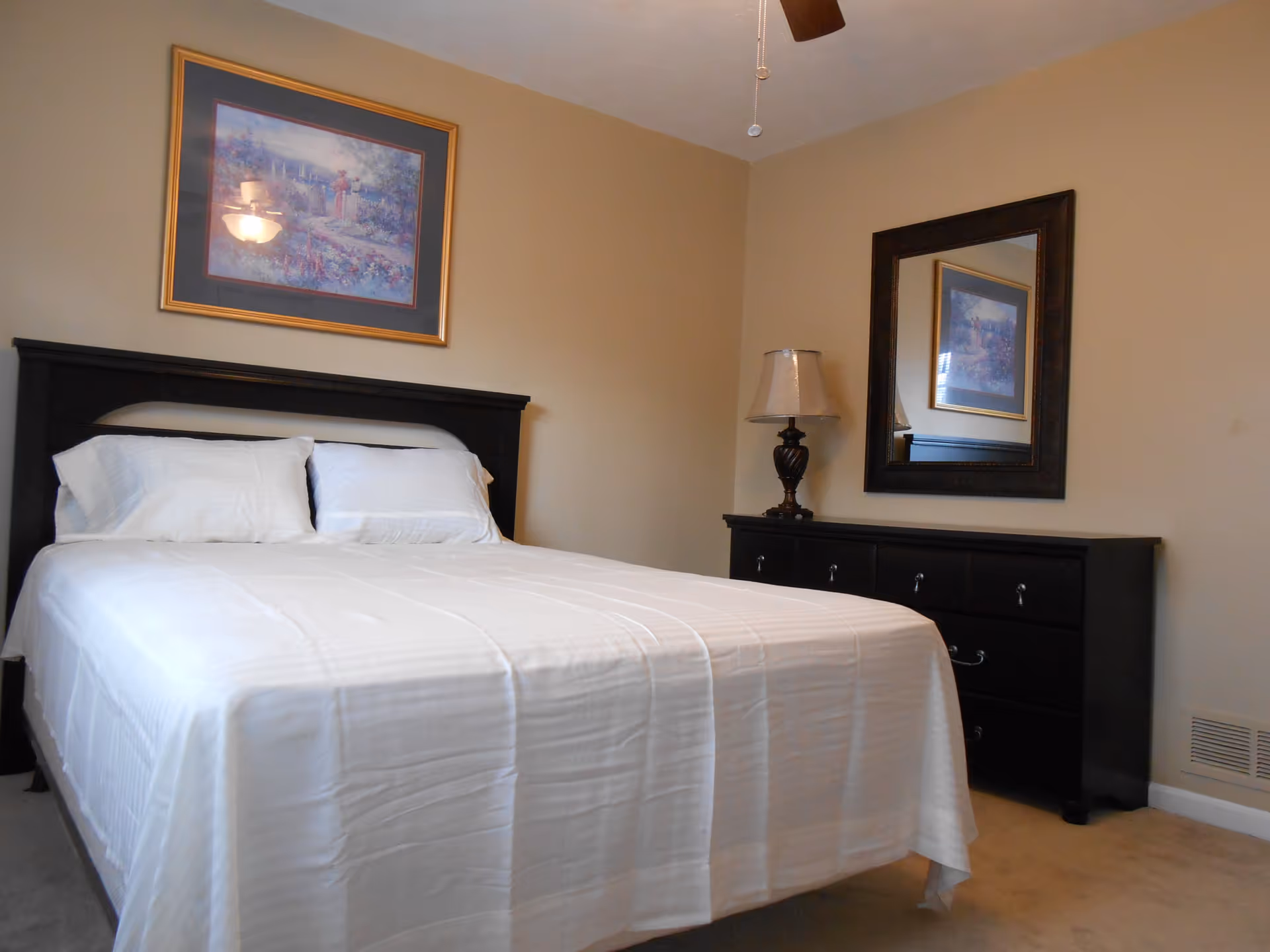 A bedroom with a neatly made bed covered in white linens and two pillows. Above the bed is a framed painting. To the right of the bed is a dark wooden dresser with a table lamp and a large mirror hanging above it. The walls are painted beige and the floor is carpeted.