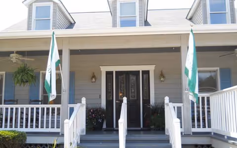 Front porch of a residential-style building with a double black door entrance, white railings, two flags on poles, hanging plants, and dormer windows on the roof.