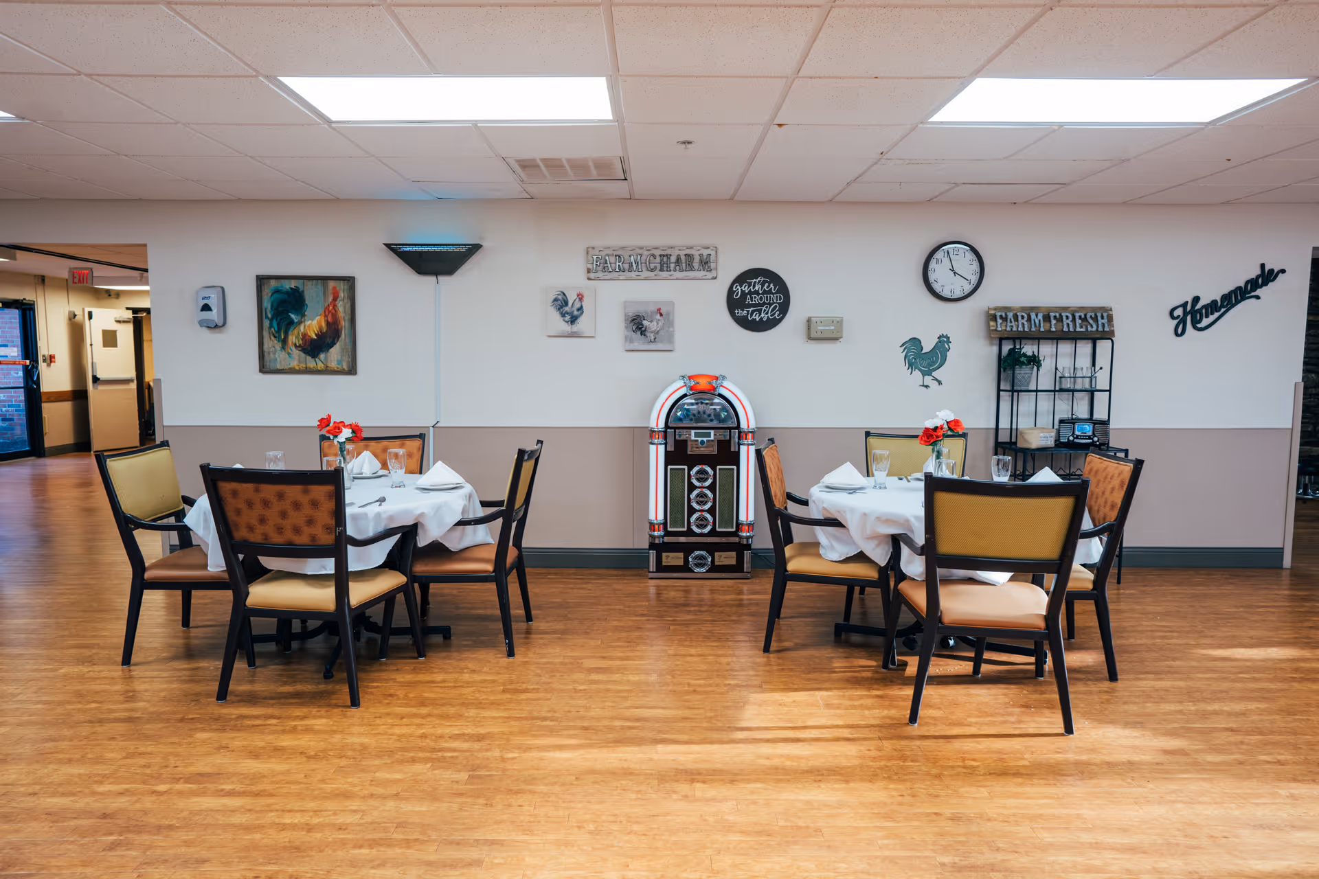 A dining area with two round tables each set with white tablecloths, napkins, glasses, and small flower arrangements. The room has wooden flooring and white walls decorated with rooster-themed artwork, a clock, and signs that say 'Farm Charm', 'Farm Fresh', and 'Homemade'. A jukebox is positioned between the tables against the wall.
