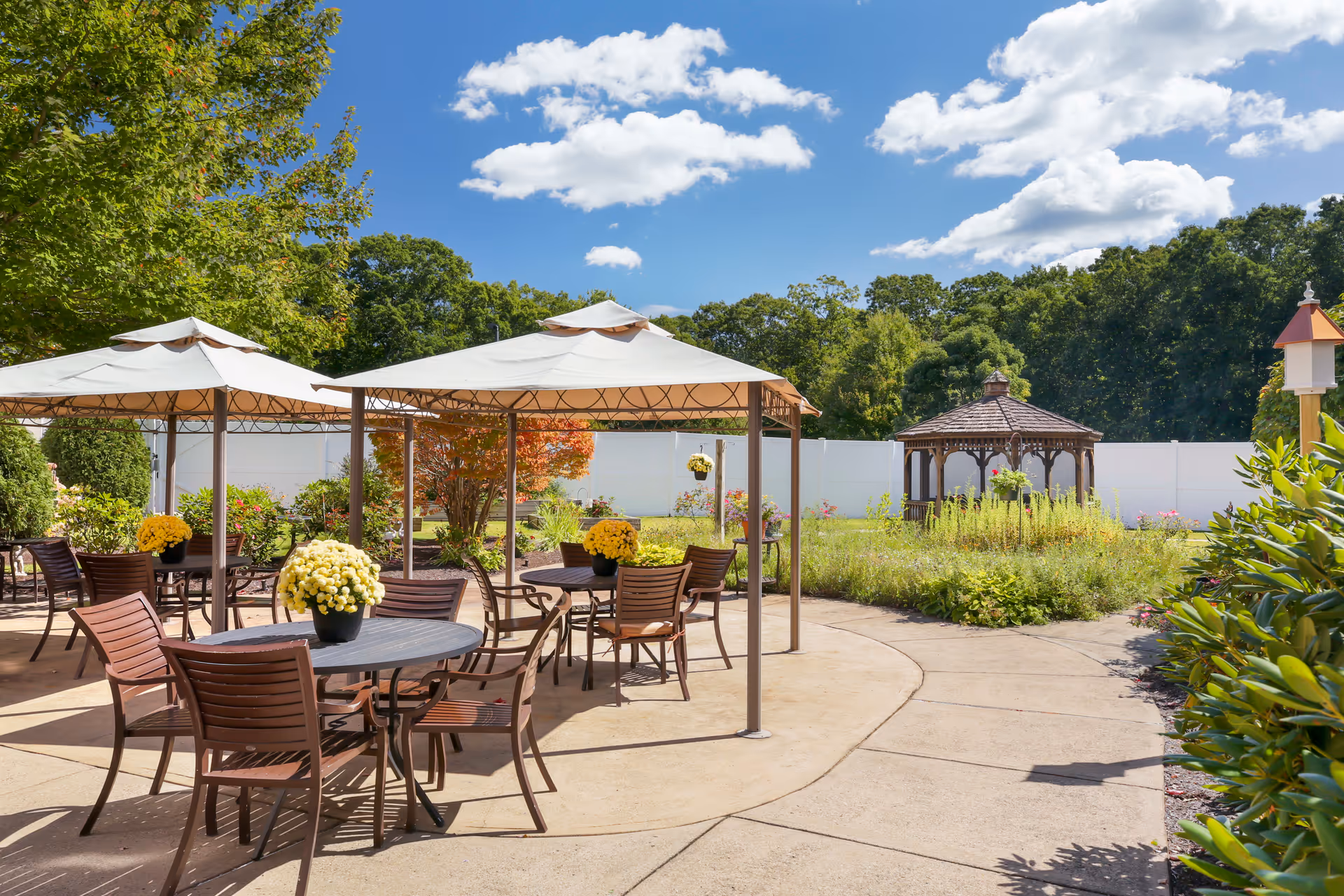 Outdoor patio area with several tables and chairs under beige canopies, decorated with potted yellow flowers. In the background, there is a wooden gazebo surrounded by greenery and a white fence, under a blue sky with scattered clouds.