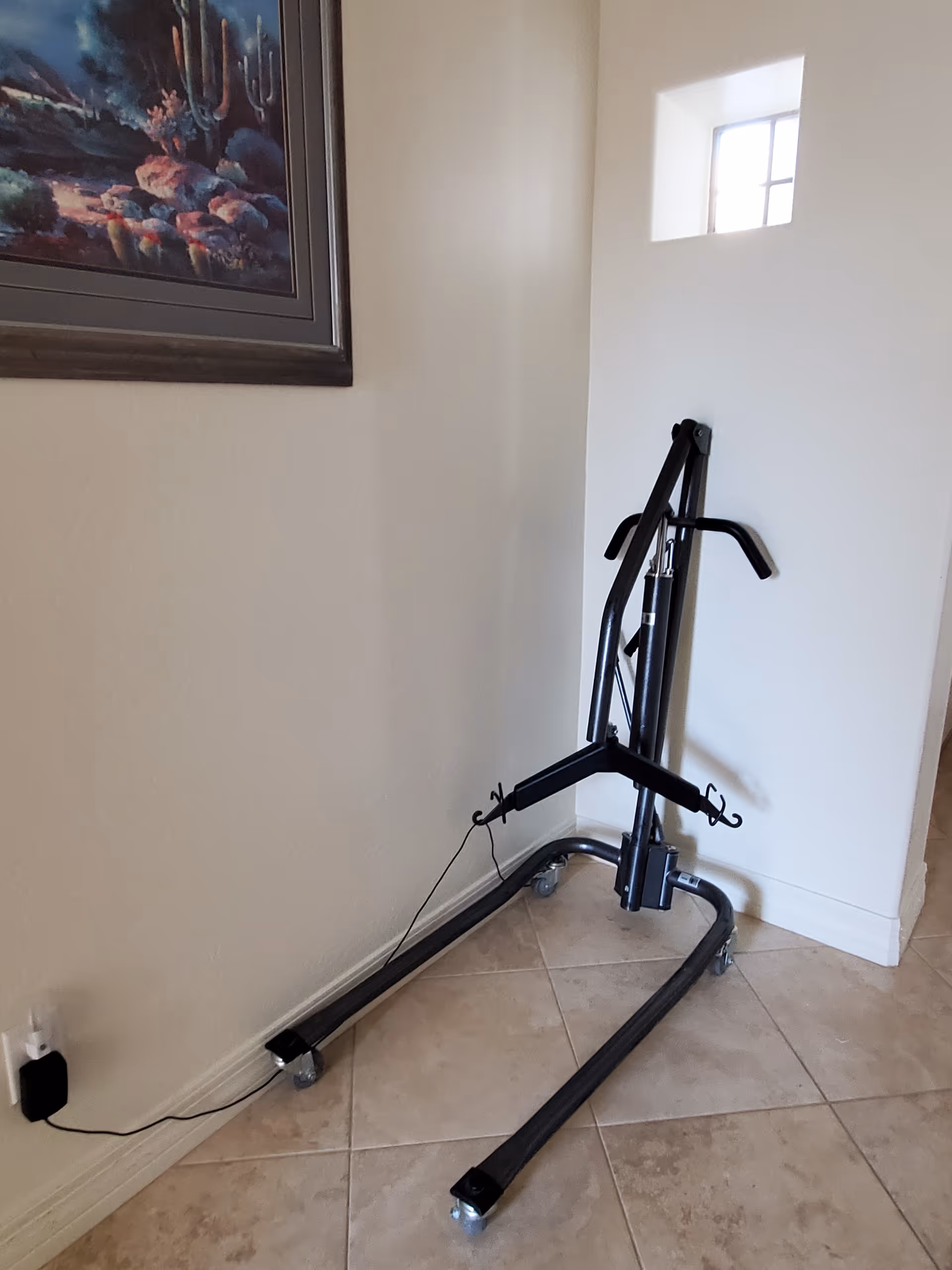 A black metal patient lift device with wheels is positioned in the corner of a tiled floor room with beige walls. There is a small window on the wall and a framed painting of a desert landscape with cacti hanging on the adjacent wall.