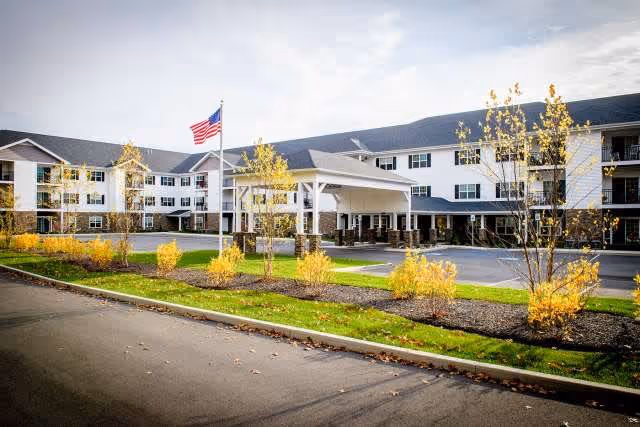 Exterior view of a large, three-story senior living facility with white siding and dark roofing. The building features multiple windows and a covered entrance with a driveway. There are young trees with yellow leaves planted along the front, and an American flag is flying on a flagpole near the entrance.