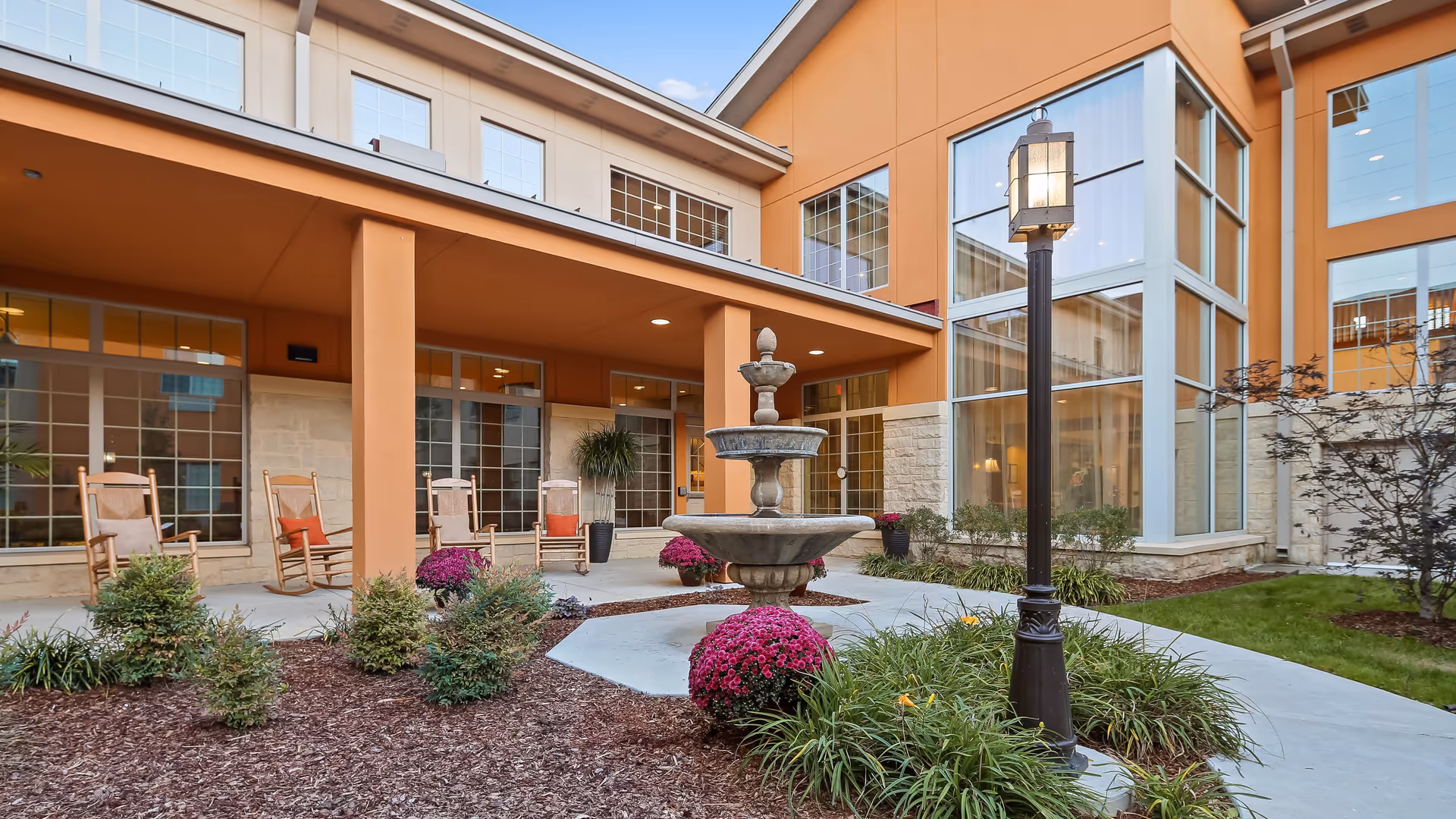 Outdoor courtyard area of a senior living facility with a central tiered water fountain surrounded by landscaping and flowers. There are several wooden rocking chairs with cushions placed under a covered patio attached to the building. The building has large windows and a warm orange exterior with stone accents. A vintage-style lamp post is also visible near the fountain.