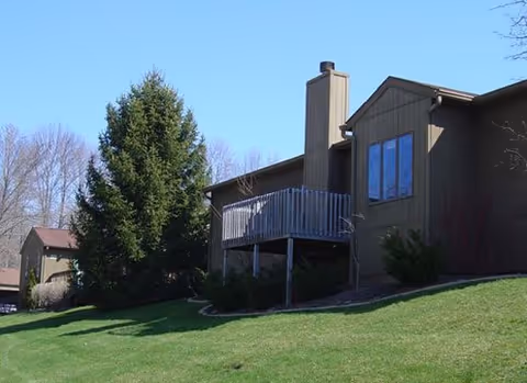 Brown single-story residential building with a raised wooden deck, green lawn, and trees under a clear blue sky.