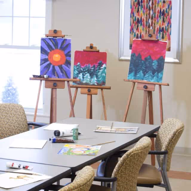 A room with a table and chairs arranged for an art activity. On the table are art supplies including paint tubes, brushes, and paper. In the background, three easels display colorful paintings featuring abstract and nature-inspired designs. The room has large windows letting in natural light and a framed artwork on the wall.