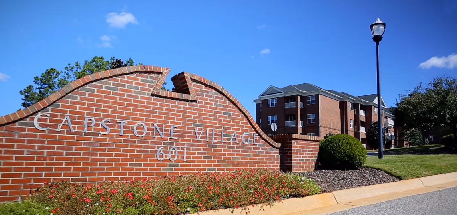Brick entrance sign for Capstone Village with the number 601, surrounded by landscaping with flowers and bushes. In the background, there is a multi-story brick building and a street lamp under a clear blue sky.