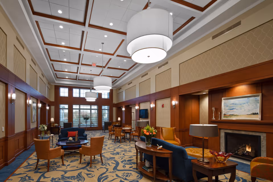 A spacious and elegant living room area in Fellowship Village featuring high ceilings with recessed lighting and large hanging lamps. The room is furnished with comfortable chairs, sofas, and tables arranged around a fireplace with a painting above it. Large windows at the far end allow natural light to fill the space, and the room has a patterned carpet and wood-paneled walls.