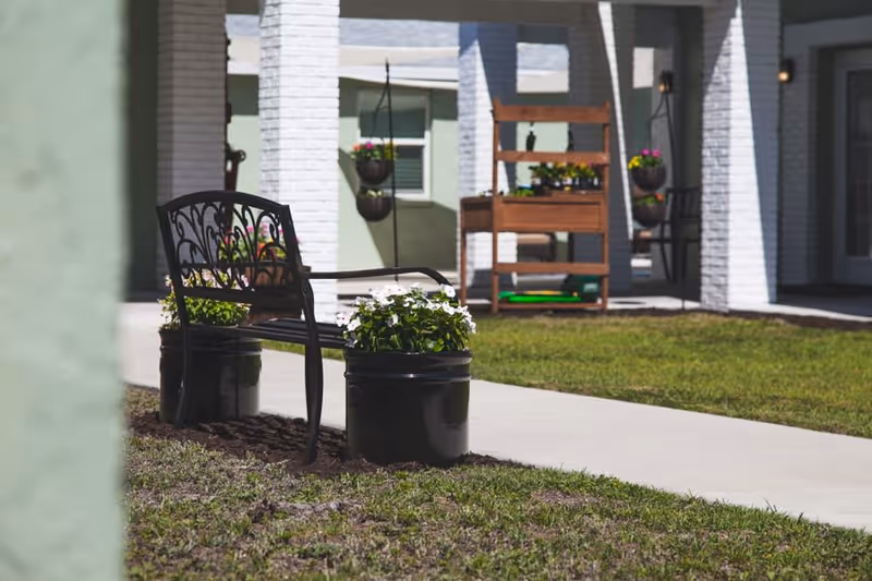 Outdoor seating area with a decorative metal bench flanked by two black planters with white flowers, situated on a grassy lawn near a paved walkway. In the background, there are white brick columns, hanging flower pots, and a wooden plant stand with potted plants, all part of a building exterior.