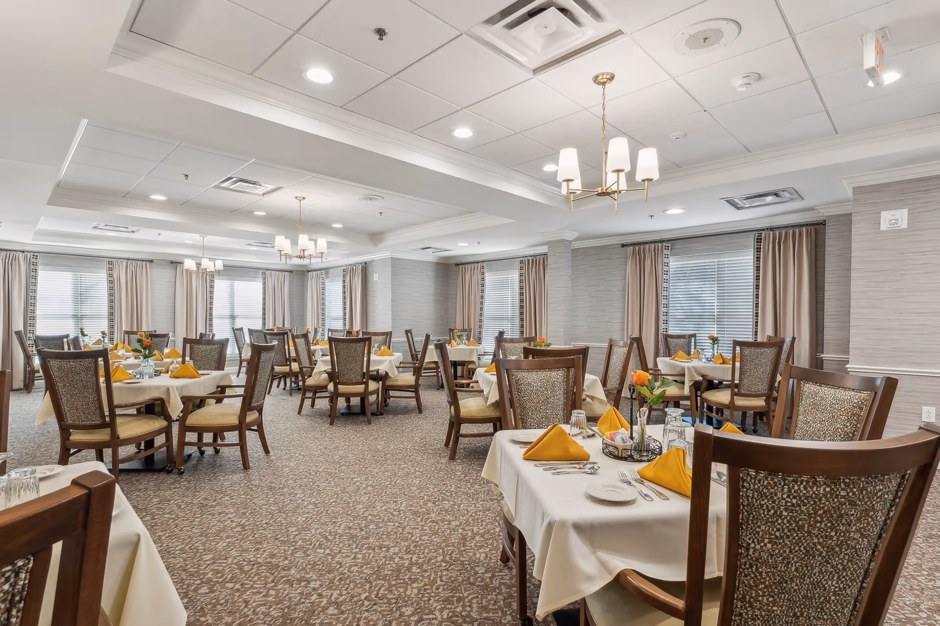Spacious dining room with multiple round tables set with yellow napkins, wooden chairs, chandeliers, and large windows with curtains.
