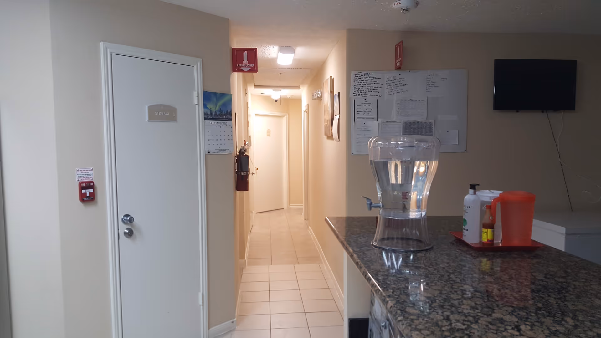 Interior hallway and reception counter showing a water dispenser and sanitizer on a granite countertop, a wall-mounted TV, and doors down the corridor.