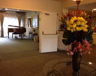 Interior common area with a large floral arrangement in a vase in the foreground and open double doors revealing a room with a grand piano.