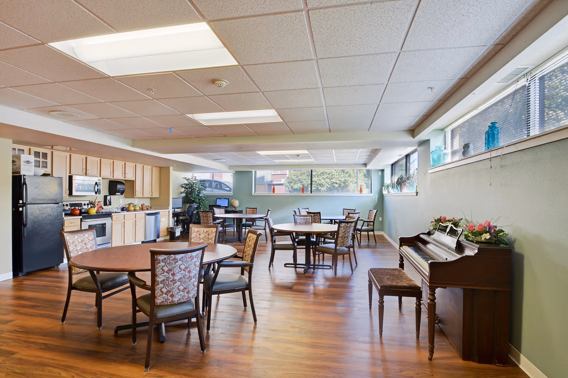 A bright and spacious common area with several round tables and chairs arranged on a wooden floor. On the left side, there is a kitchen area with light wood cabinets, a black refrigerator, microwave, stove, and dishwasher. On the right side, there is a piano with a bench and floral decorations on top. Large windows along the back and right walls let in natural light, and decorative items are placed on the window sills.