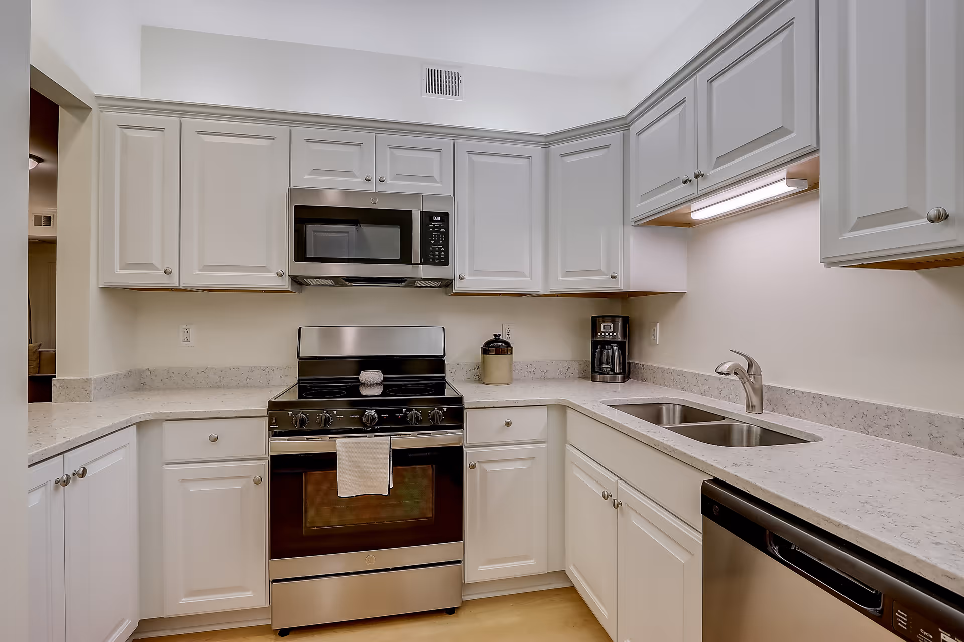 A modern kitchen with white cabinetry, a stainless steel stove with oven, a microwave above the stove, a double sink, a dishwasher, and a coffee maker on the countertop. The countertops are light-colored with a subtle pattern, and there is under-cabinet lighting above the sink area.