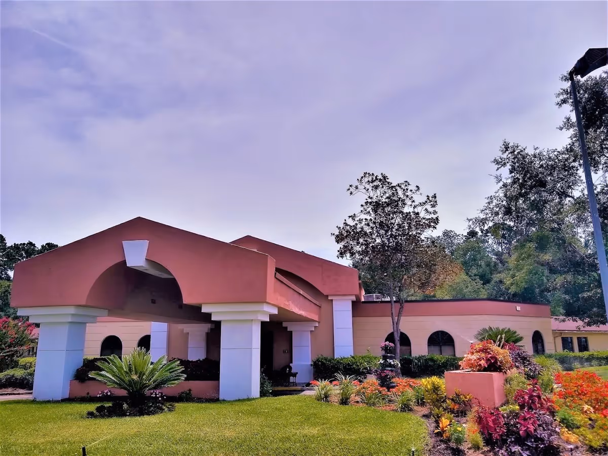 Front entrance of a pink/terracotta assisted living building with columns, landscaped lawn, and trees.