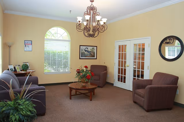 A cozy living room with beige walls and carpeted floor, featuring a chandelier hanging from the ceiling. The room contains two brown armchairs and a matching sofa arranged around a round wooden coffee table with a floral centerpiece. There is a large arched window with white blinds, a framed picture of a bird on the wall, a round mirror, and a small side table with a plant and picture frames.