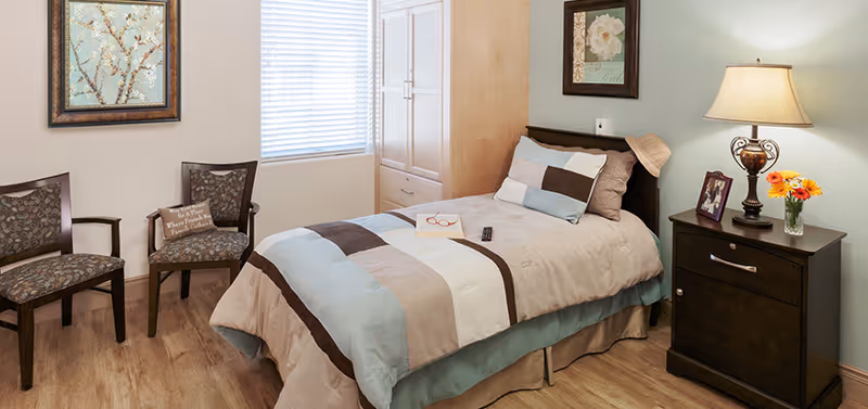 A cozy bedroom with a single bed covered in a beige, brown, and light blue striped comforter. On the bed are a pair of red reading glasses, a book, and a TV remote. Next to the bed is a dark wooden nightstand with a lamp, a framed photo, and a small vase of orange and yellow flowers. Two patterned chairs are placed against the wall near a window with blinds. The walls are decorated with framed floral artwork, and the floor is wooden.