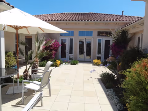 Outdoor patio area with tiled flooring, white chairs, a glass table, and a large white umbrella. The patio is surrounded by plants and flowers, with a building featuring multiple glass doors and windows in the background under a red-tiled roof.