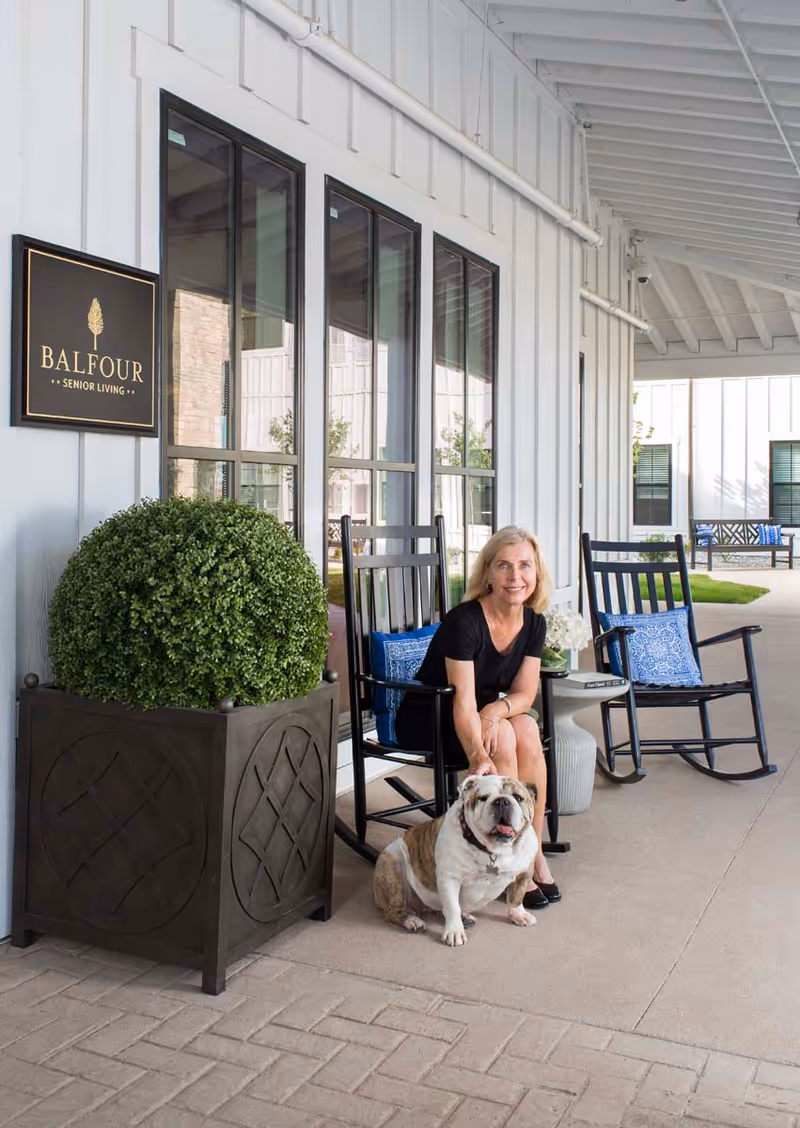 A woman sitting on a black rocking chair on a covered patio area outside a senior living facility named Balfour Central Park. She is petting a bulldog sitting on the ground in front of her. The patio has large windows, another empty rocking chair with a blue cushion, a small table with flowers, and a large decorative planter with greenery.