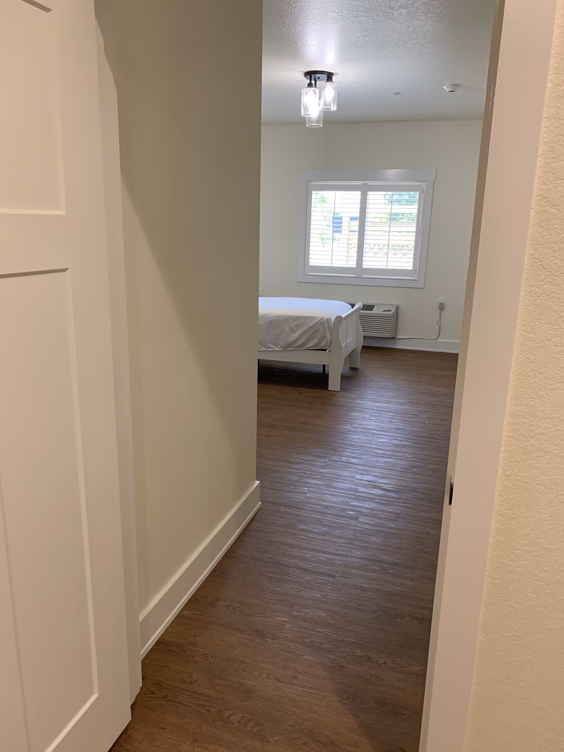 View down a short hallway into a simple bedroom with a white bed, window shutters, wood-look flooring and a ceiling light.