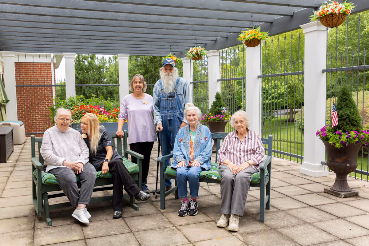 A group of five elderly people and one caregiver sitting and standing on a covered outdoor patio area with hanging flower baskets and potted plants. The patio has a tiled floor and white columns with a black metal fence, overlooking a green garden area.