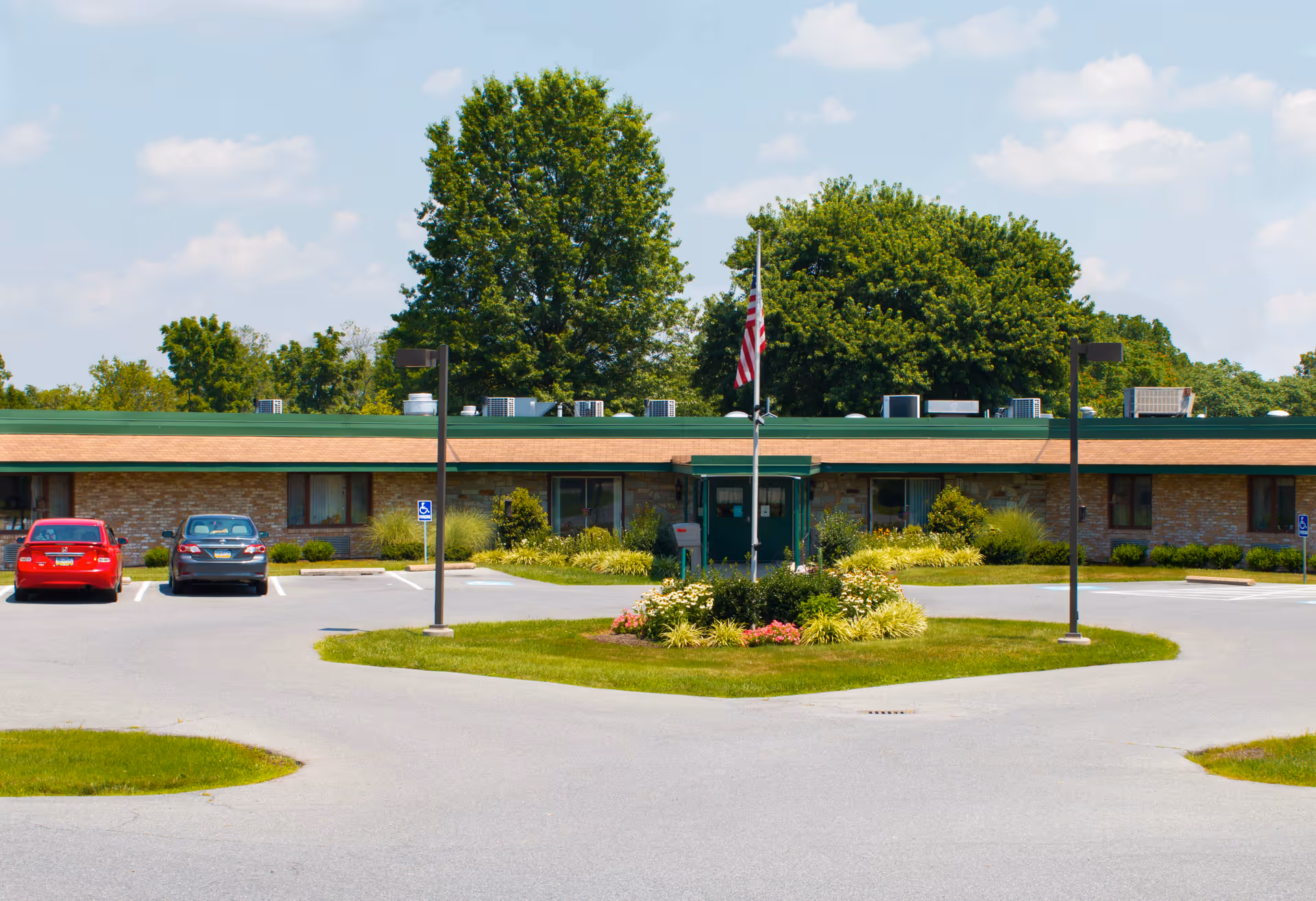 Single-story brick building with a green roof, surrounded by a parking lot with two cars parked and a landscaped circular island with an American flag in the center. Large trees are visible behind the building under a partly cloudy sky.