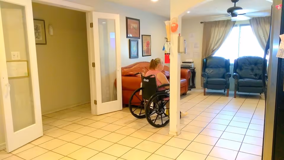 A woman in a wheelchair is sitting in a living room area with tiled floors. The room has a brown leather couch, two dark armchairs near a window with curtains, framed artwork on the walls, and a ceiling fan. There is a white pillar in the middle of the room and double doors leading to another area.