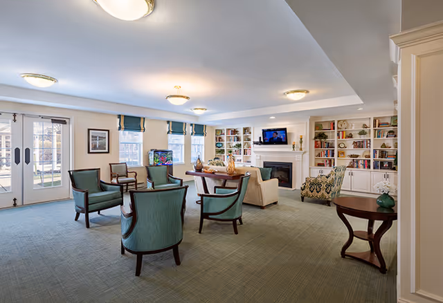 A spacious senior living common area with green carpet, several green upholstered chairs arranged around a wooden table, a beige armchair, a patterned armchair, built-in white bookshelves filled with books and decorative items, a fireplace with a TV mounted above it, and large windows with blue valances letting in natural light.