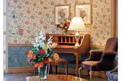 Small seating area with floral wallpaper, a wooden desk with a lamp and framed pictures, upholstered chairs and a coffee table topped with a vase of flowers.