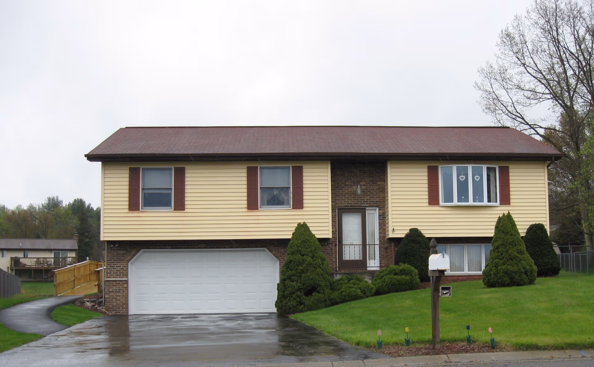 Front exterior view of a two-story house with a brown roof, yellow siding, and brick lower walls. The house has a white garage door, three windows with red shutters, a front door with a small porch, and a green lawn with bushes and a mailbox.