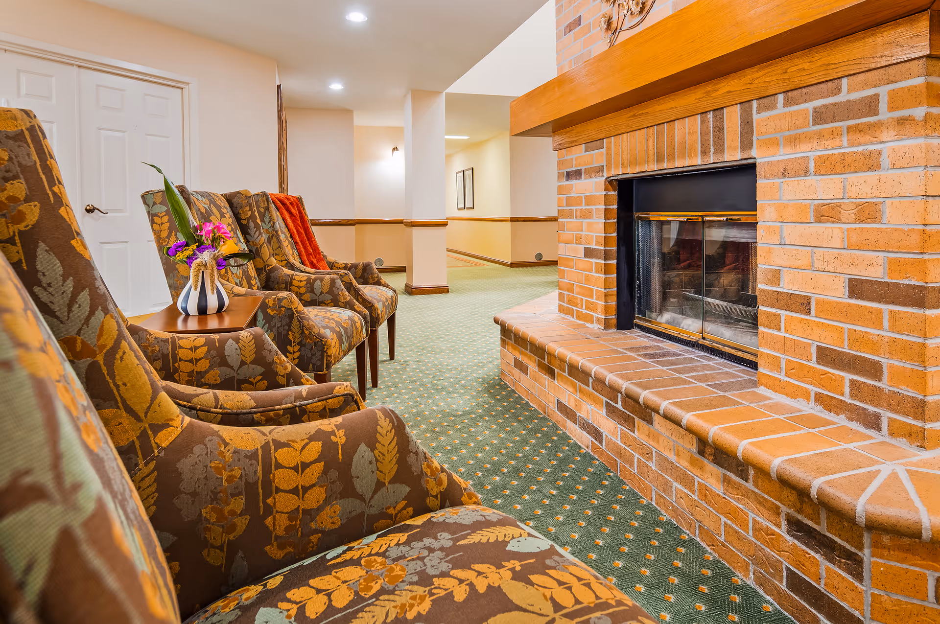 Row of patterned armchairs arranged beside a brick fireplace in a carpeted common area.