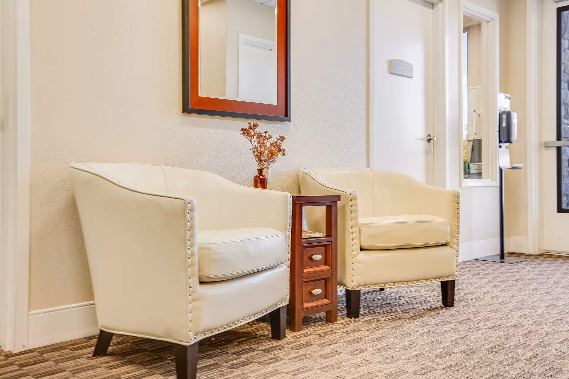 Two cream leather armchairs with a small wooden side table and vase under a wall mirror in a hallway seating area.