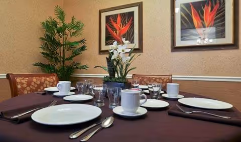 A dining table set with white plates, cups, glasses, and silverware on a dark tablecloth. In the center of the table is a floral arrangement with white flowers. The background shows a beige wall with two framed pictures of red and orange flowers and a green potted plant.