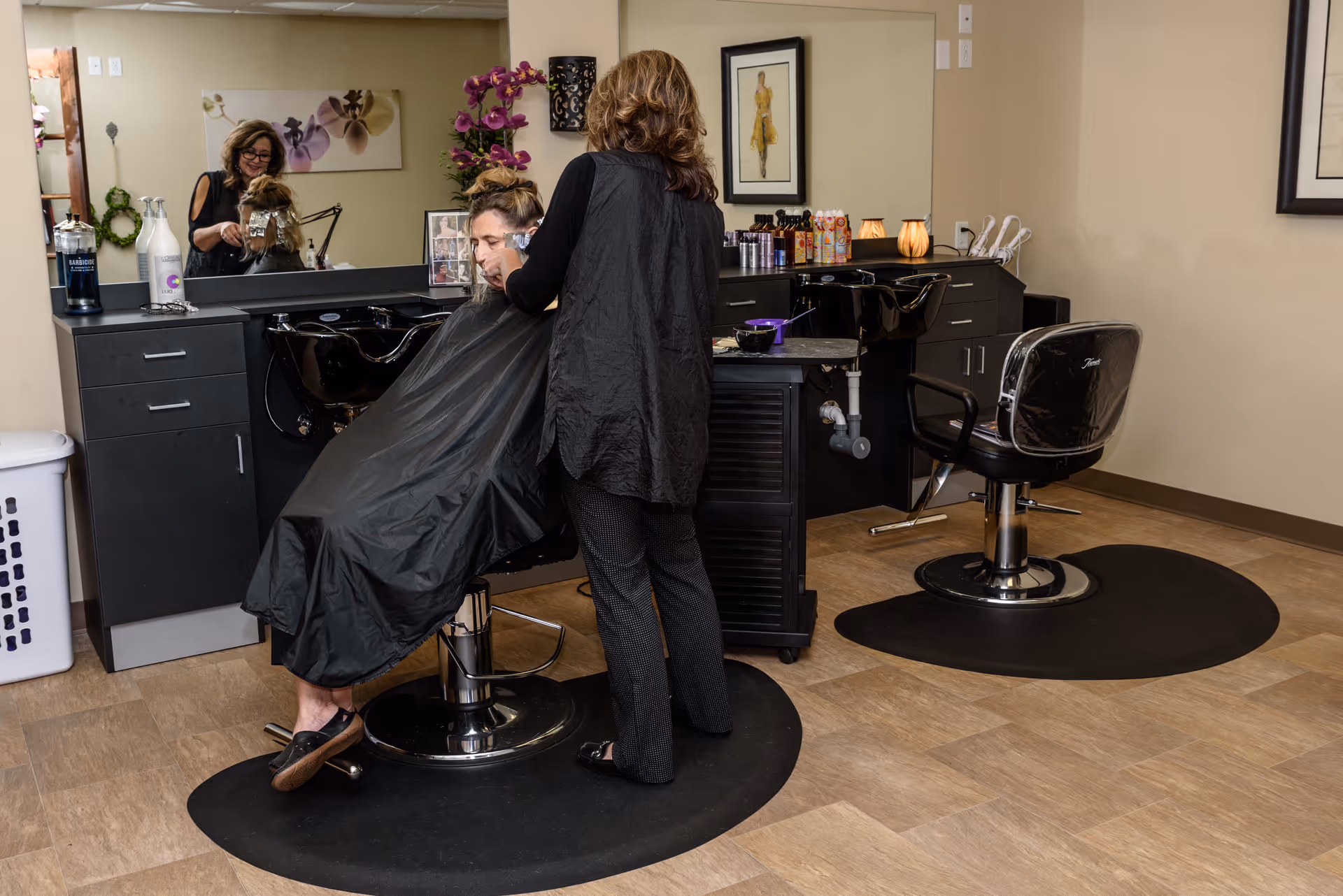 A hair salon area inside Oak Park Retirement Resort with a stylist applying hair treatment to a seated woman. The room features black salon chairs, sinks for washing hair, a large mirror, and various hair care products on the counter. The floor is covered with wood-patterned tiles and black mats under the chairs.
