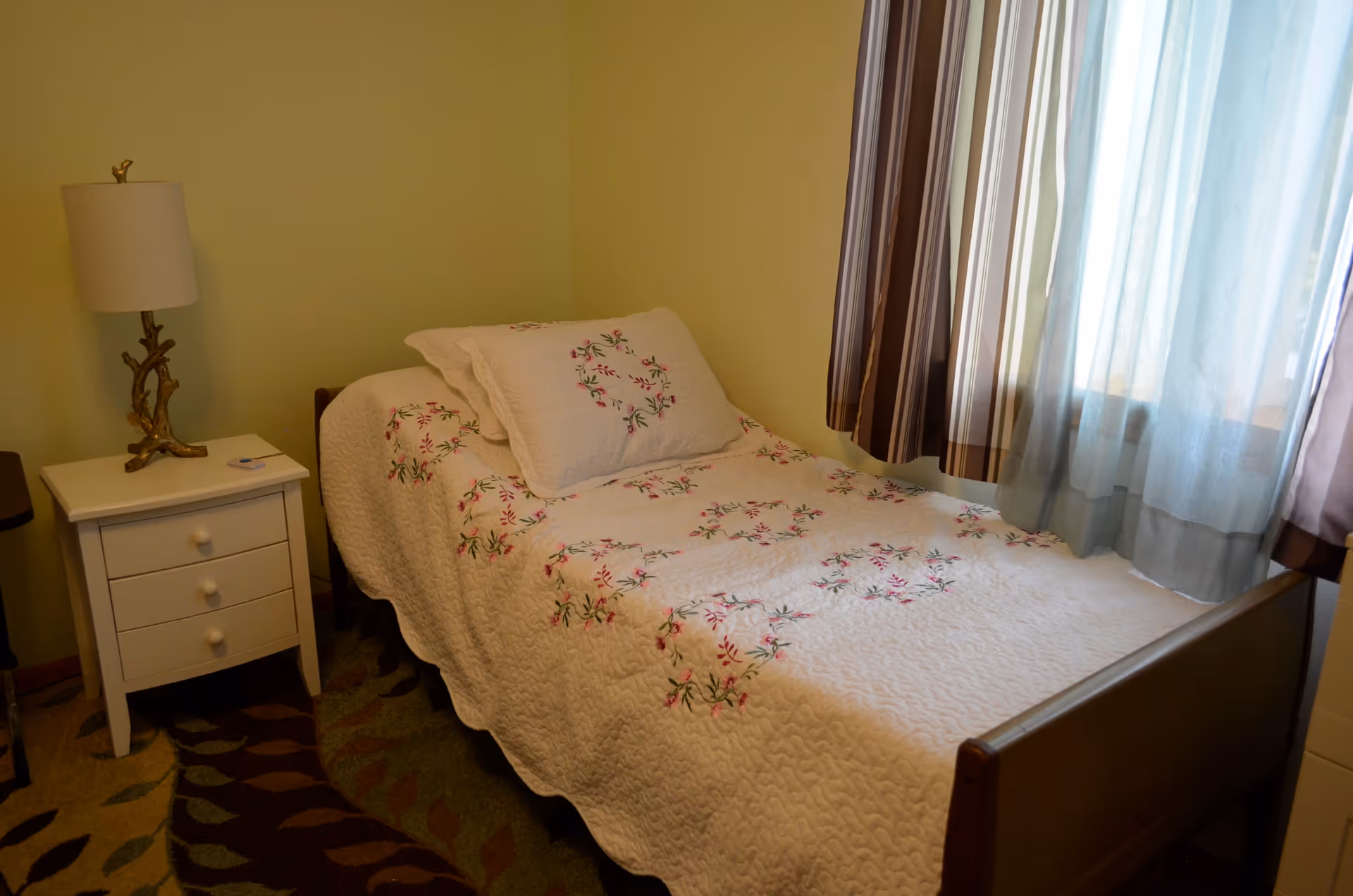 A small bedroom with a single bed covered in a white quilt with floral embroidery. Next to the bed is a white nightstand with three drawers and a decorative lamp. A window with sheer and striped curtains allows natural light into the room.