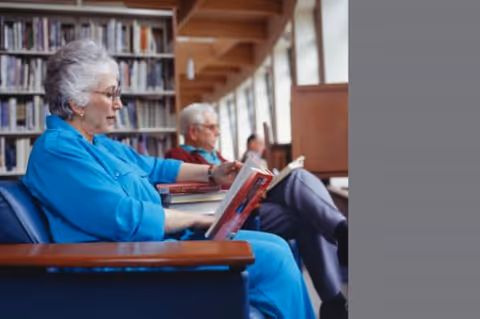 An elderly woman in a blue outfit sitting on a blue chair reading a book in a library or reading room, with bookshelves in the background. An elderly man is seated behind her, also reading.