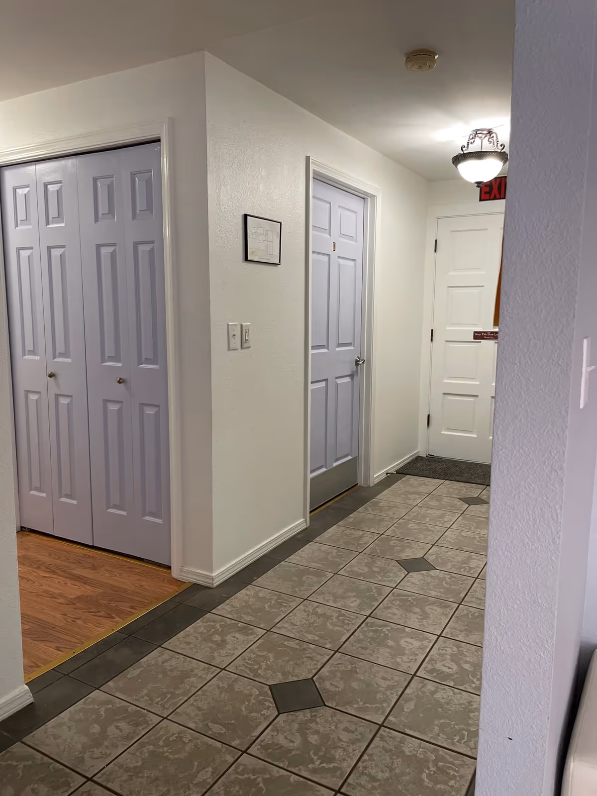 Interior hallway with tiled floor and white walls. There are three closed doors visible, two painted light purple and one white with an exit sign above it. A framed floor plan is mounted on the wall, and a ceiling light fixture is also visible.