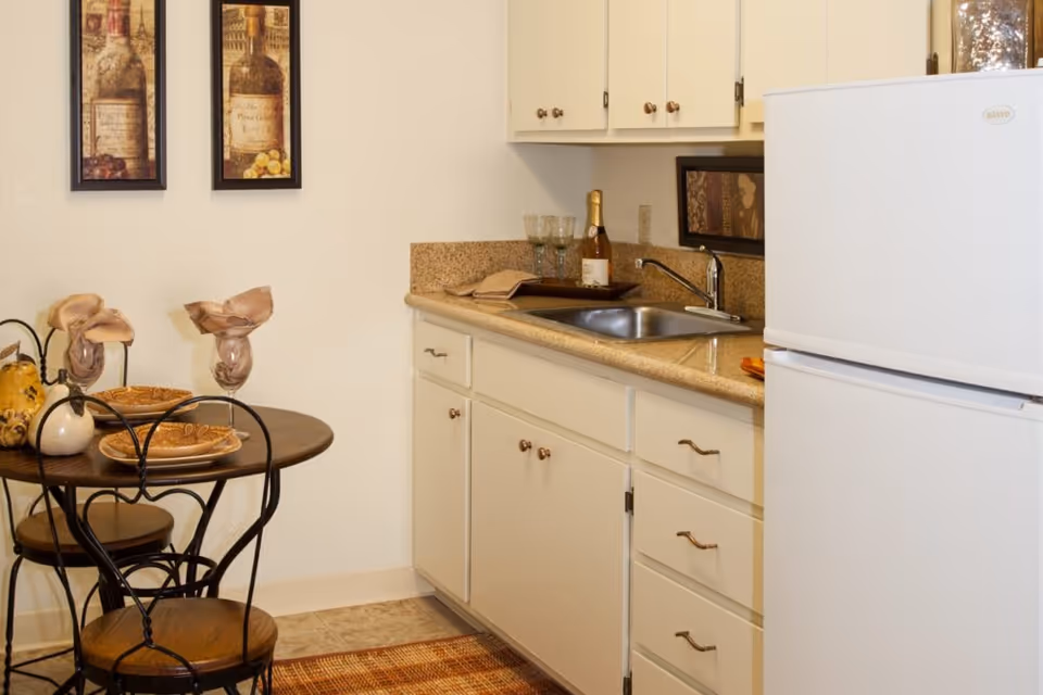 A small kitchen area with white cabinets, a granite countertop, a stainless steel sink, and a white refrigerator. Next to the kitchen is a small round wooden table set with two plates, two glasses with napkins, and decorative items. Two framed pictures of wine bottles hang on the wall above the table.