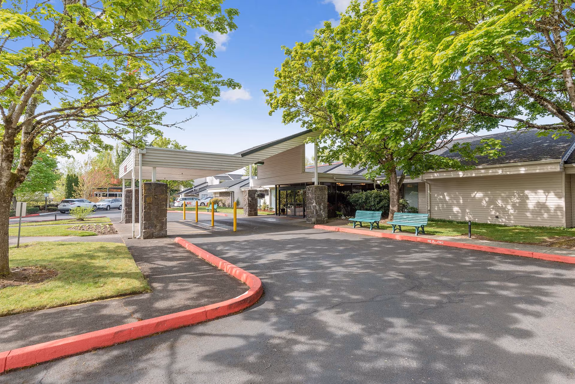 Entrance driveway of Holiday Summerfield Estates with a covered drop-off area supported by stone pillars, surrounded by green trees and benches on the right side, and parked cars visible in the background under a clear blue sky.
