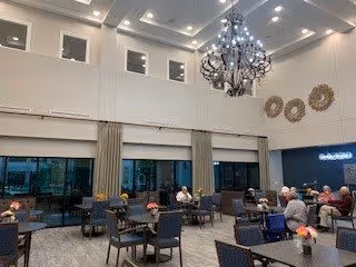 A spacious dining room with high ceilings and large windows. Several tables with chairs are arranged throughout the room, with people seated and engaged in conversation. The room features modern lighting, including a large chandelier, and decorative wall art. Curtains hang beside the windows, and the overall atmosphere is bright and welcoming.