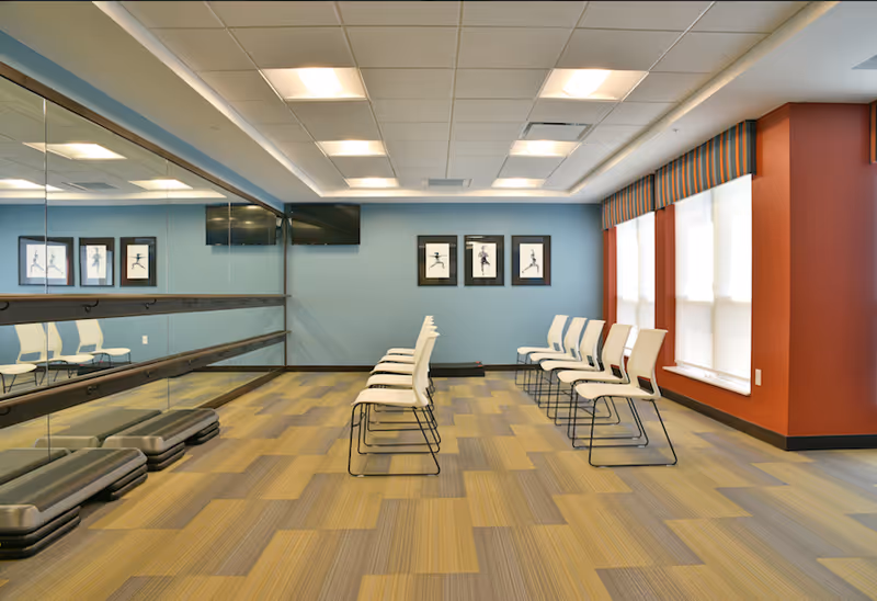 Bright multipurpose exercise room with rows of white chairs facing a mirrored wall, step platforms, framed artwork, and large windows.