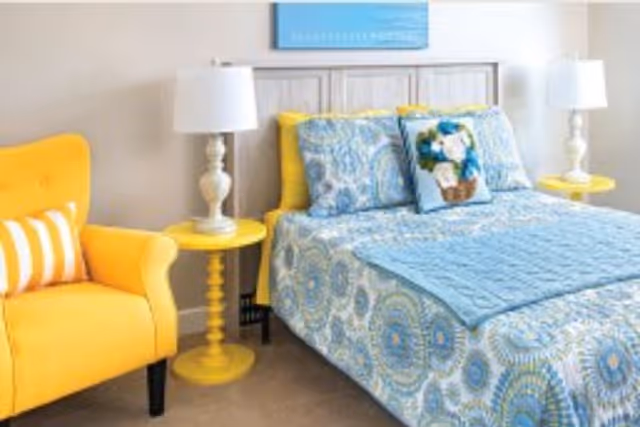 Bright bedroom with blue patterned bedding, a wooden headboard, yellow side tables and lamps, and a yellow armchair.