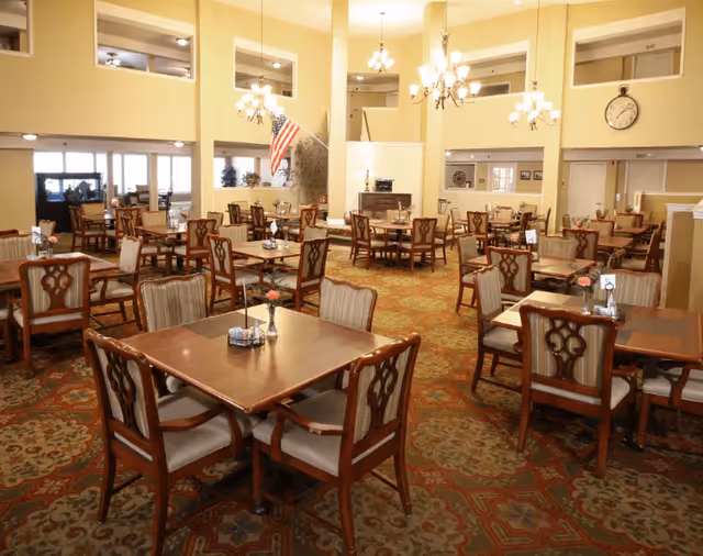 A spacious dining room with multiple wooden tables and chairs arranged neatly on a patterned carpet. The room features high ceilings with chandeliers, large windows, an American flag, and a clock on the wall. The atmosphere is warm and inviting, suitable for communal dining.
