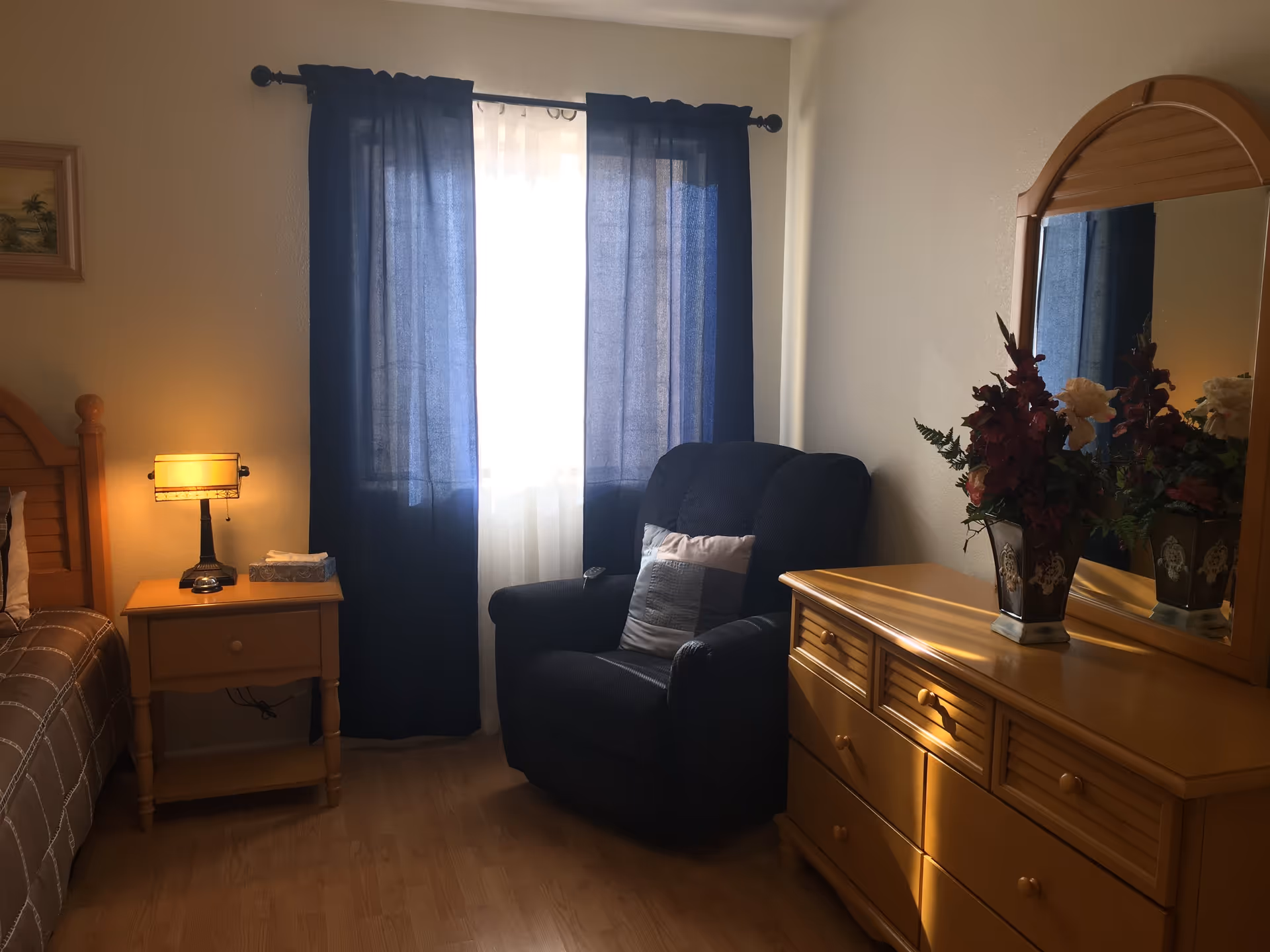 A cozy bedroom with a wooden bed partially visible on the left, a wooden nightstand with a lamp and a box of tissues, a dark blue armchair with a cushion in front of a window with blue curtains, and a wooden dresser with a mirror and a vase of flowers on top.