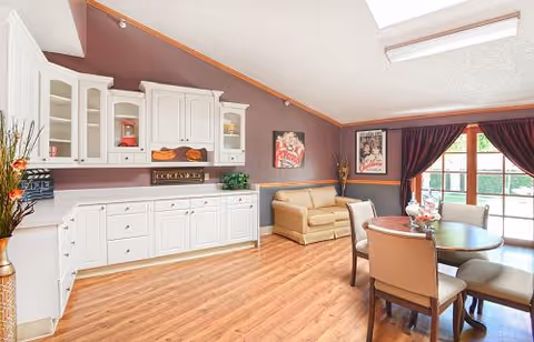 Bright communal dining and sitting area with a round table and chairs, a tan sofa, white cabinetry, and wood floors.